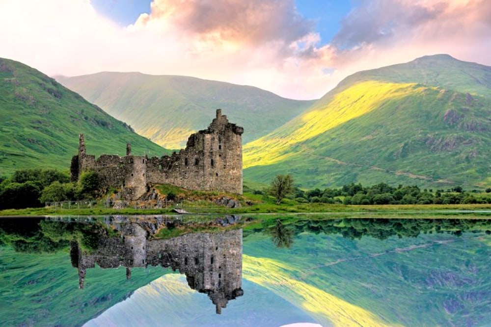 A ruined castle on a lochside, mirrored in calm water below green hills